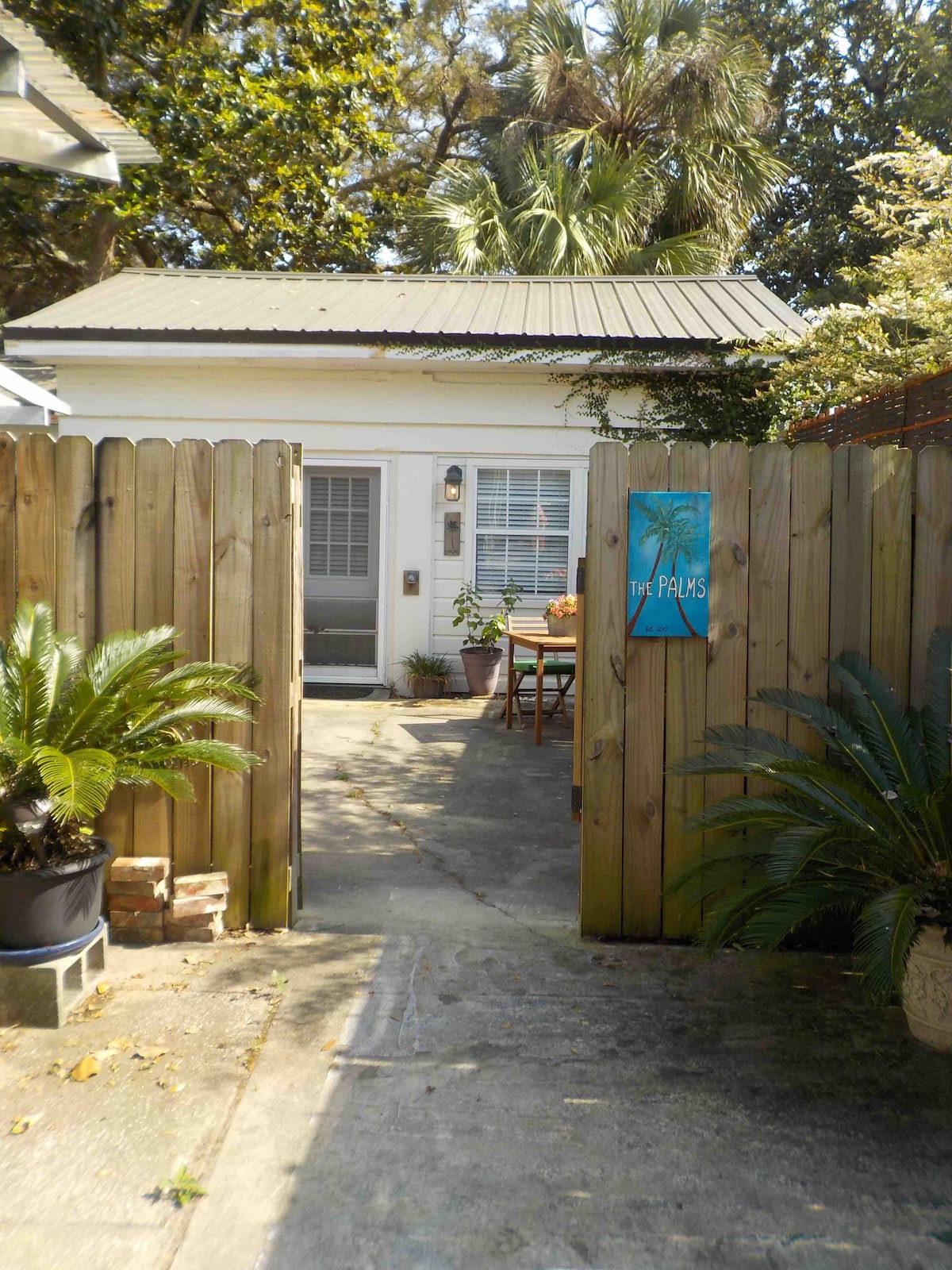 An inviting entrance is framed by a wooden fence and lush greenery. A pathway leads to the cottage, with a small table and chairs positioned to the side. A painted sign labeled 'The Palms' adds a colorful touch to the scene.
