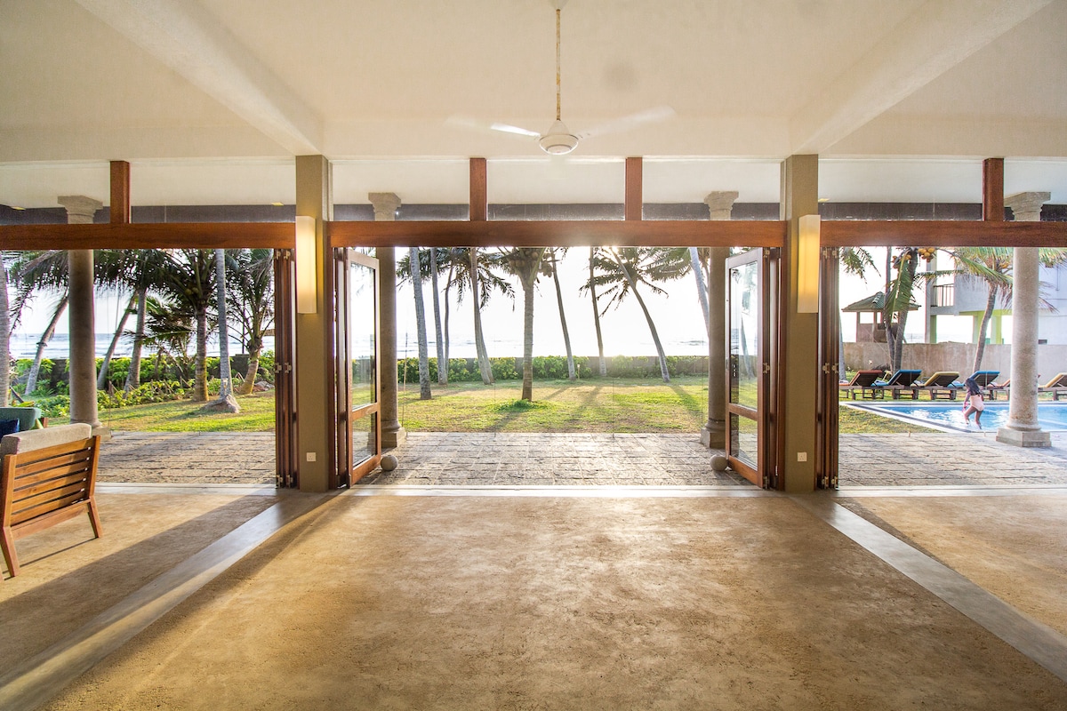 A spacious indoor area opens to the garden and beach, framed by large glass doors. Natural light floods the space, highlighting the smooth, neutral flooring. The inviting view includes swaying palm trees, a serene lawn, and a glimpse of the pool in the distance.
