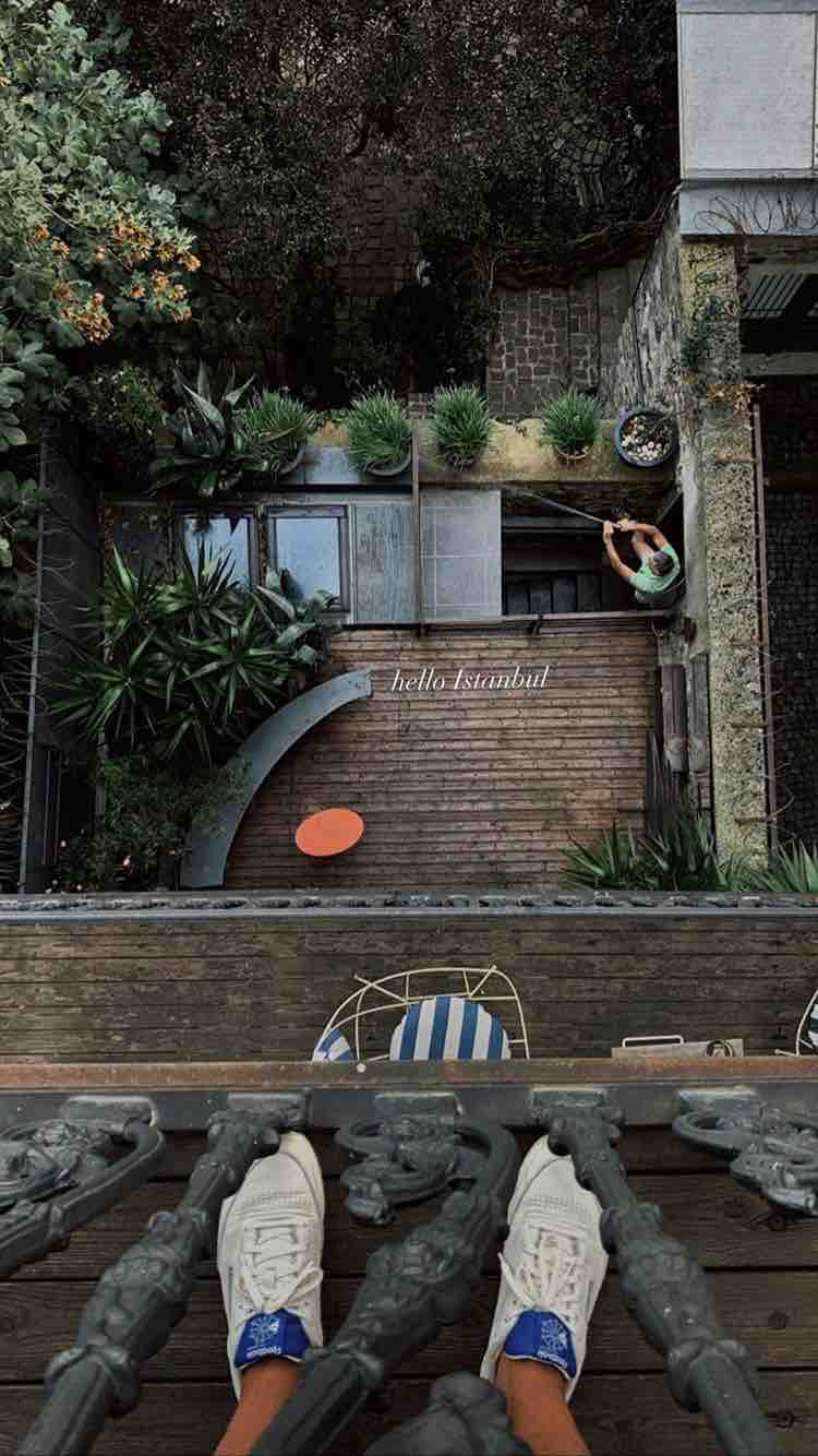 An aerial view reveals a backyard adorned with lush greenery and various plants. A curved wooden path leads to a seating area with striped chairs, while a person tends to the garden. The scene is framed by the balcony's ornate railing.