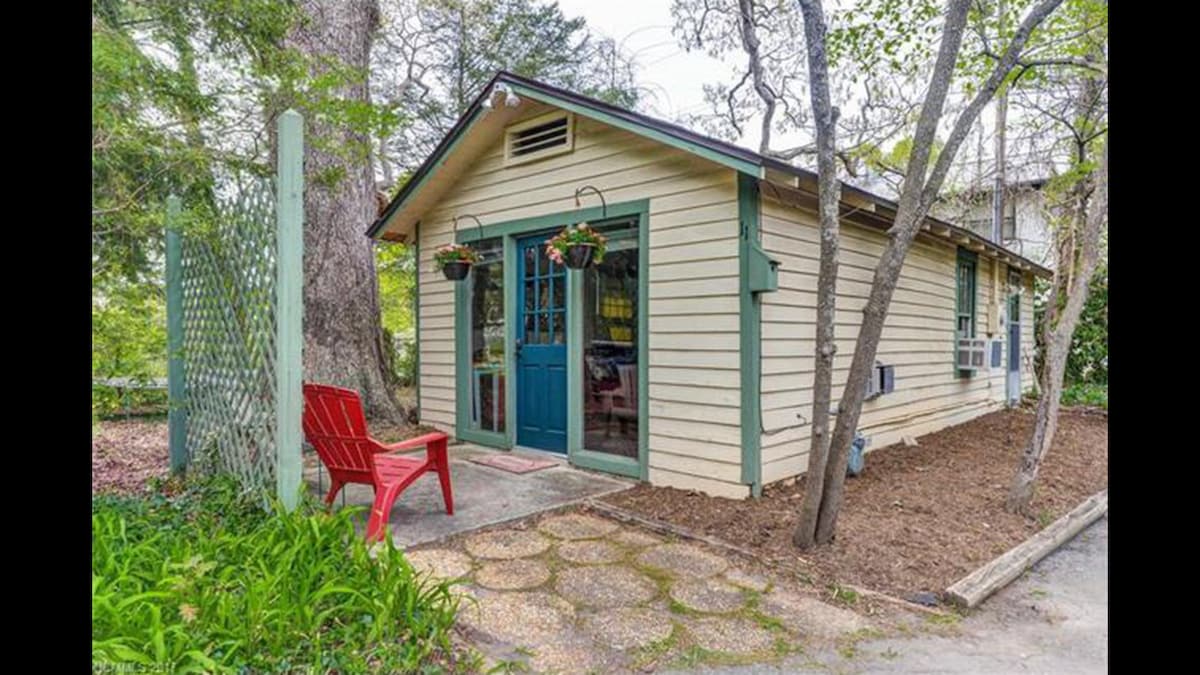 A charming exterior of a cottage is displayed with a blue door and large windows framed by green foliage. A red Adirondack chair is positioned on the path leading to the entrance, while planters hang from the porch, adding a touch of greenery to the setting.