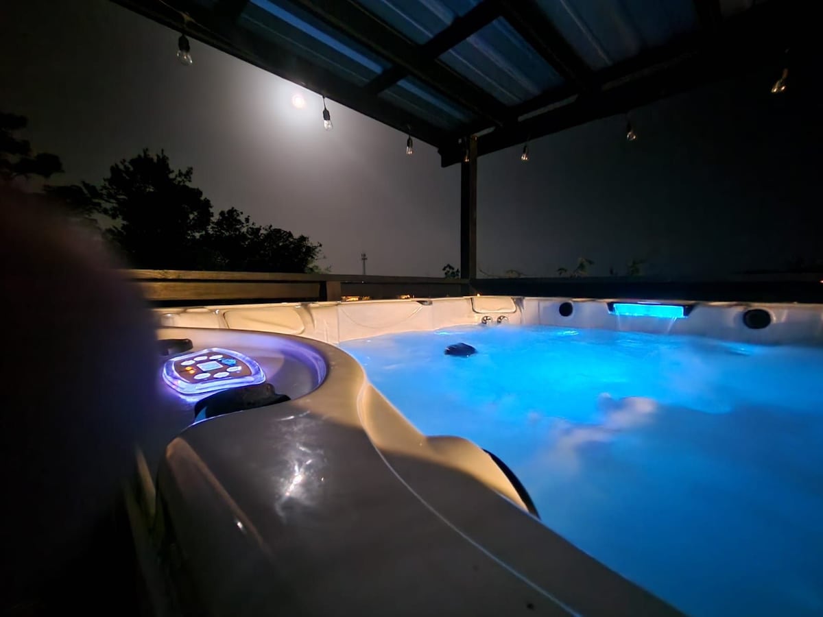 A spa Jacuzzi is illuminated with soft blue lighting, creating a calming ambiance under a night sky. The structure offers an open-air design, with views of surrounding trees in the background. A moonlit glow enhances the tranquil setting for relaxation.