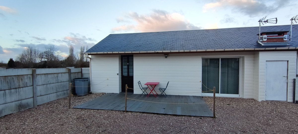 A single-story building features a wooden deck area with a small red table and two chairs. Large sliding glass doors provide access to the interior, while a gravel pathway surrounds the space. A fence and a water barrel are visible in the background.