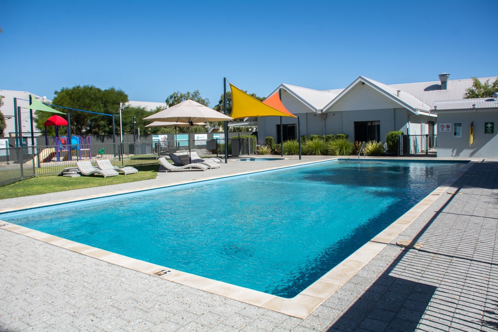 A large swimming pool is framed by a paved area and lush greenery. Sun loungers are positioned along one side, while colorful play structures are visible in a nearby area, providing a family-friendly atmosphere. A shaded seating area adds comfort for relaxation.