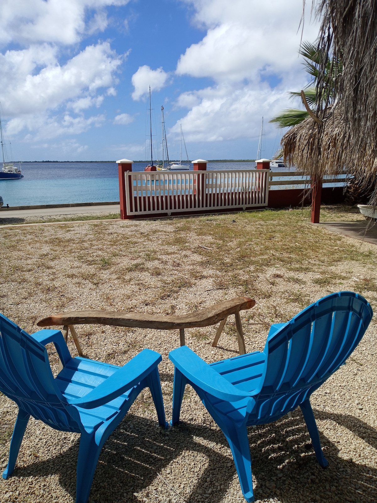 Two bright blue chairs are positioned facing the sea, creating a comfortable spot for relaxation. A wooden bench connects the chairs, while small pebbles cover the ground. Sailboats are visible in the distance against a backdrop of a partly cloudy sky.