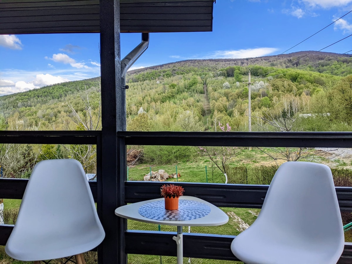 A small balcony area features two white chairs positioned around a round table, which holds a potted plant. The background displays lush green hills under a blue sky with scattered clouds, providing a serene and natural view.