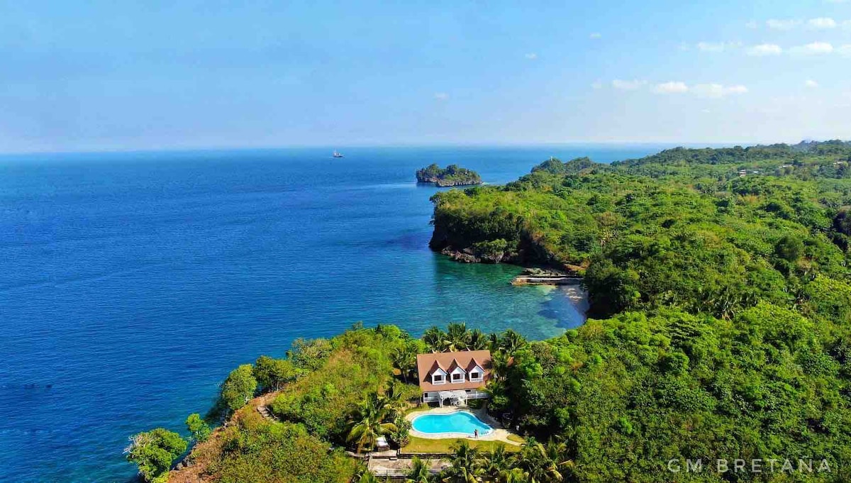 Aerial view of a lush coastal landscape showcasing a spacious private property with a swimming pool. Vibrant greenery surrounds the building, with clear blue waters gently lapping at distant cliffs. The tranquil sea extends to the horizon, enhancing the sense of privacy and natural beauty.
