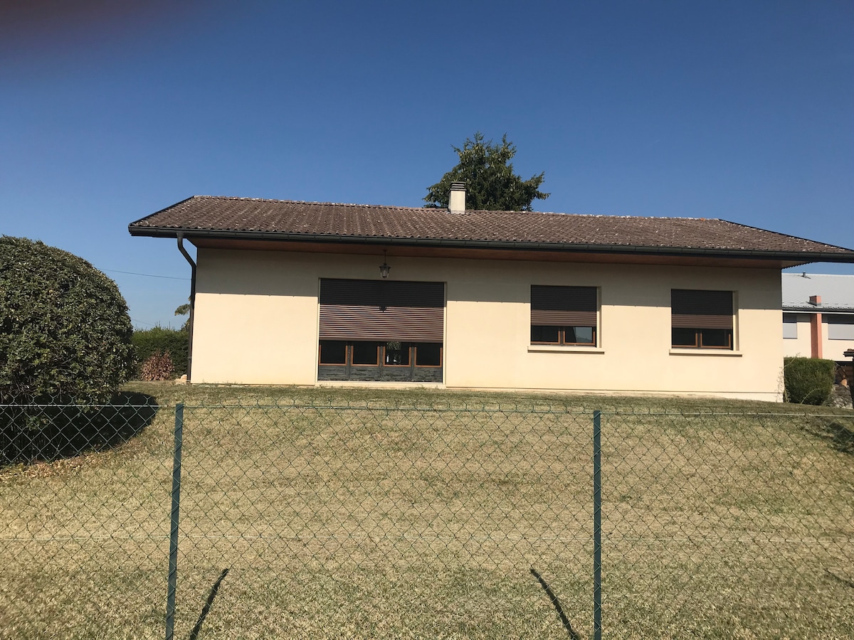 A single-story villa is set against a clear blue sky, featuring a flat roof and large windows. The surrounding garden is well-kept with a grassy lawn and is bordered by a simple fence, providing a welcoming outdoor space.