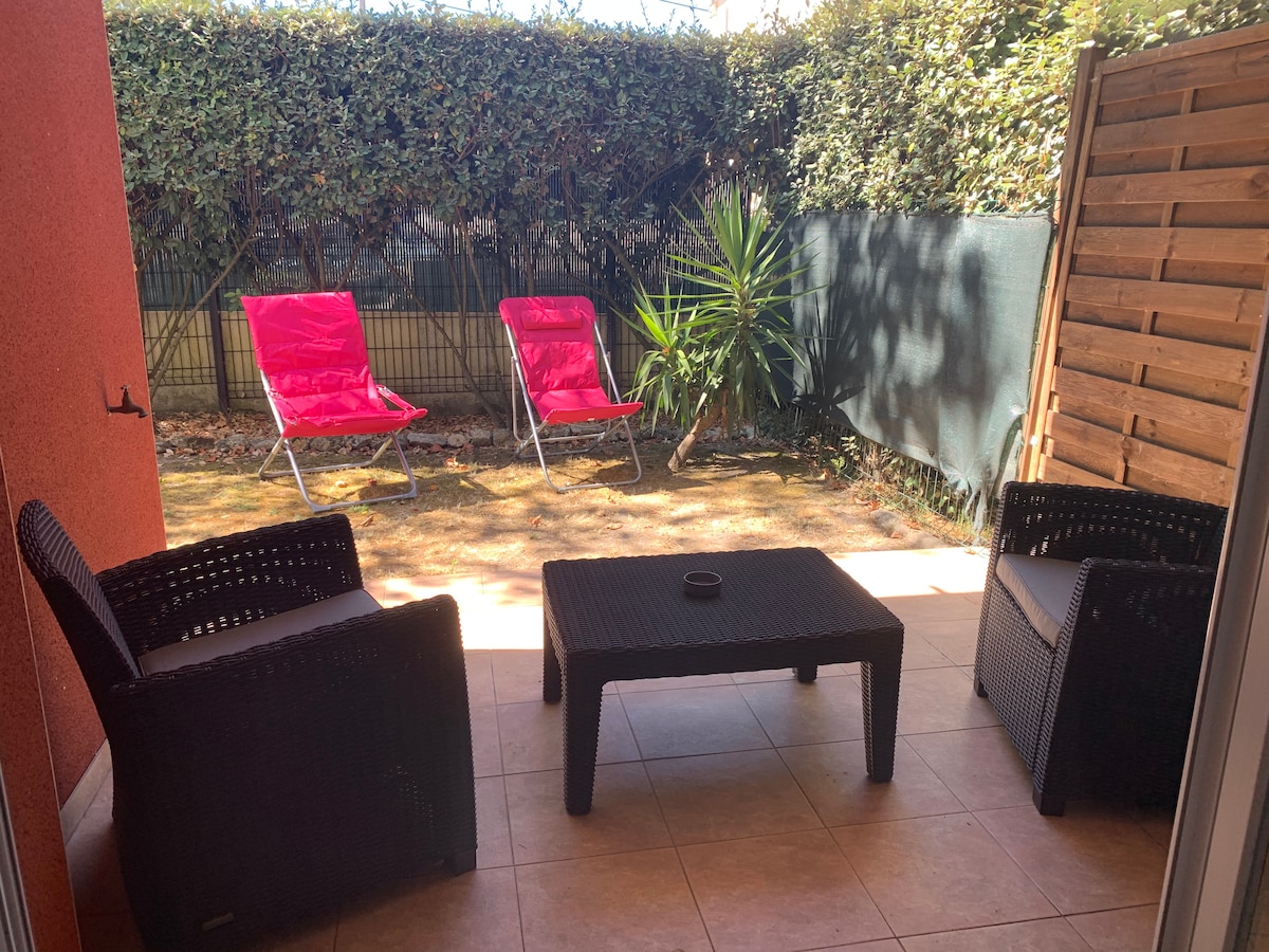 A private garden area is featured, with two bright pink lounge chairs positioned on a tiled patio. A small black coffee table is situated between the chairs, surrounded by greenery. The space is lightly shaded, creating a relaxing outdoor atmosphere.