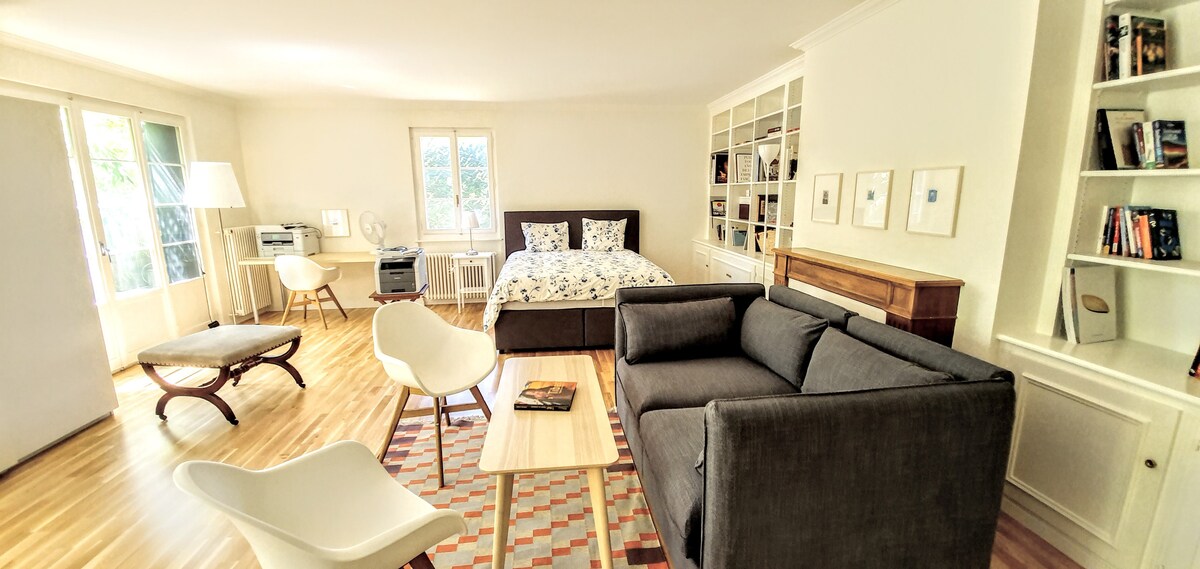 A spacious studio showcases a comfortable bed with patterned bedding. A light-filled seating area features a gray sofa, an armchair, and a coffee table. Natural light streams through large windows, illuminating the wooden flooring and decorative bookshelf filled with books.