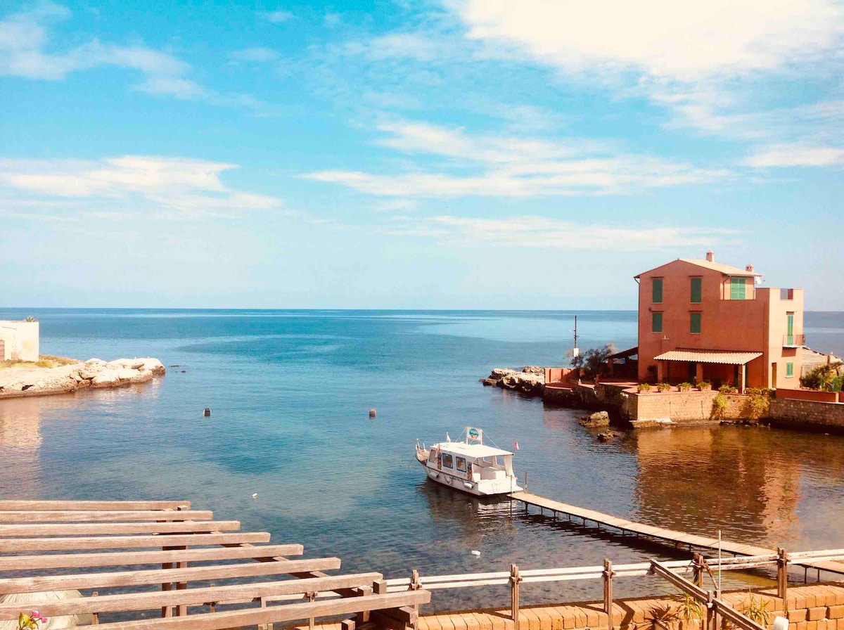 A serene coastal view reveals a calm sea reflecting blue skies. A small boat is docked near a pier, leading to a charming house with red walls, set against rocky shores. The peaceful landscape invites tranquility and offers a glimpse of coastal living.