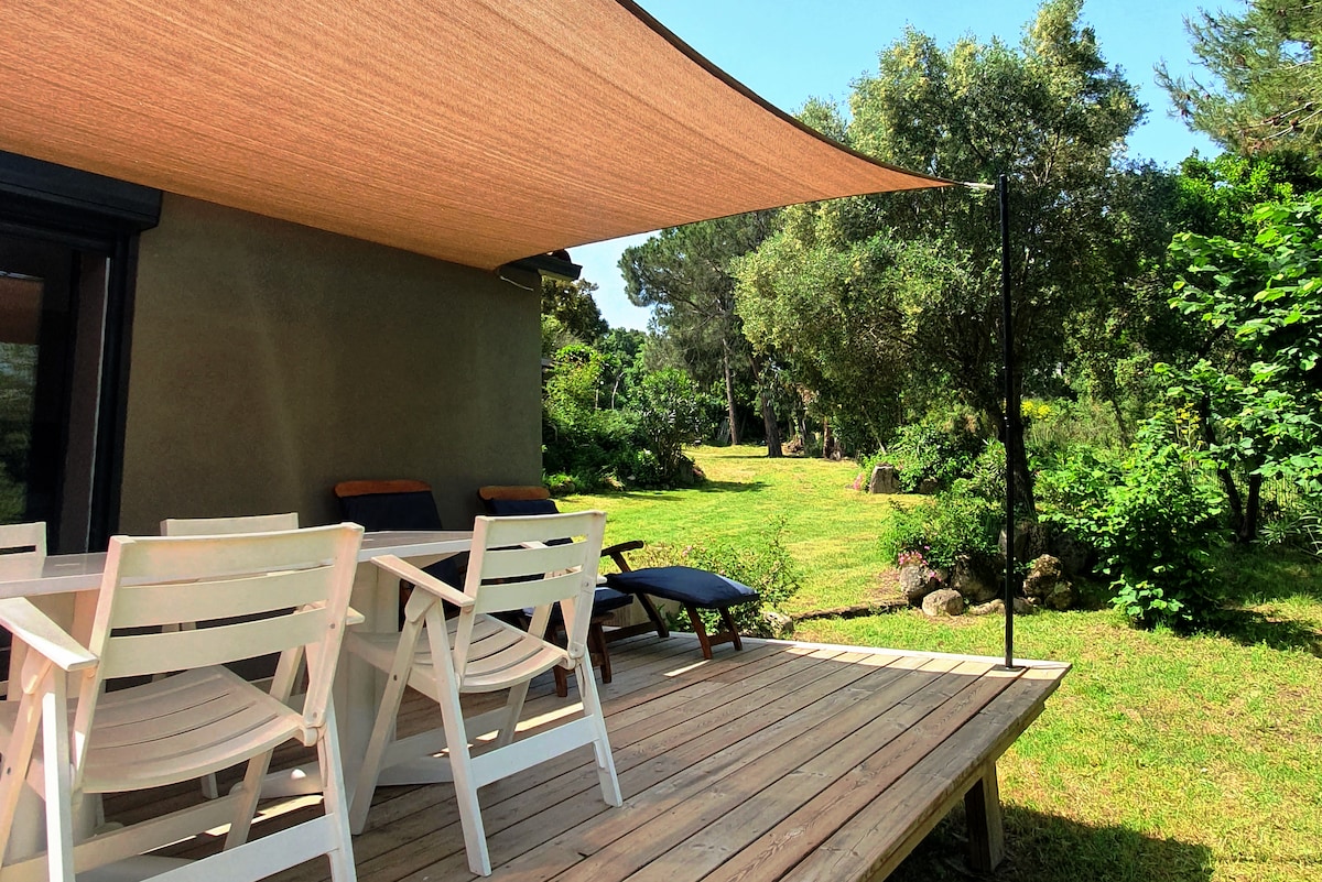 A shaded terrace features a dining table surrounded by four white chairs and a lounge chair. Lush greenery extends into the distance, enhancing the tranquil outdoor environment. The building's exterior reflects a natural color palette, blending harmoniously with the surrounding landscape.