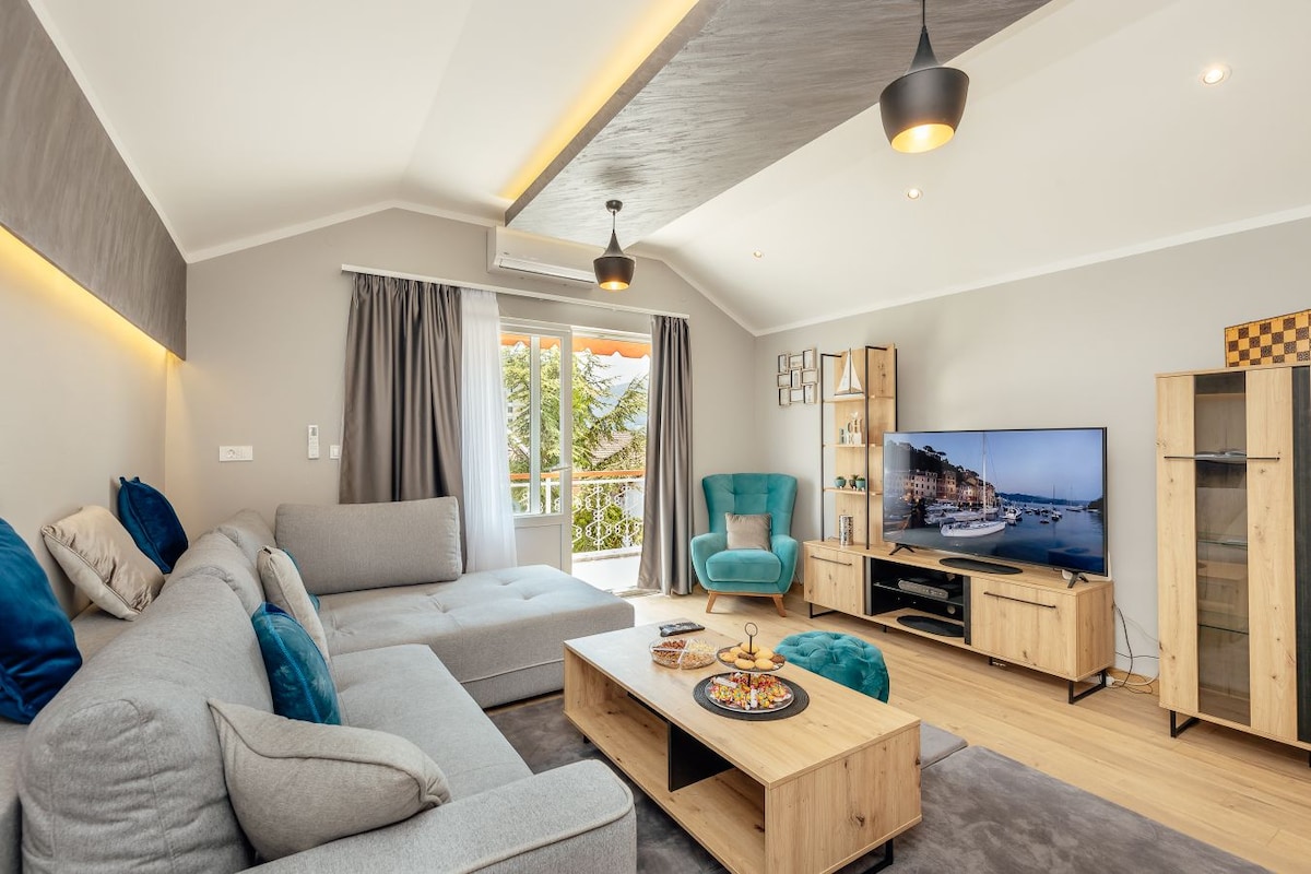 A spacious living area features a modern sectional sofa adorned with decorative cushions. Natural light enters through double doors that lead to a balcony. A television is positioned on an entertainment unit, with a wooden coffee table displaying an assortment of snacks and drinks.