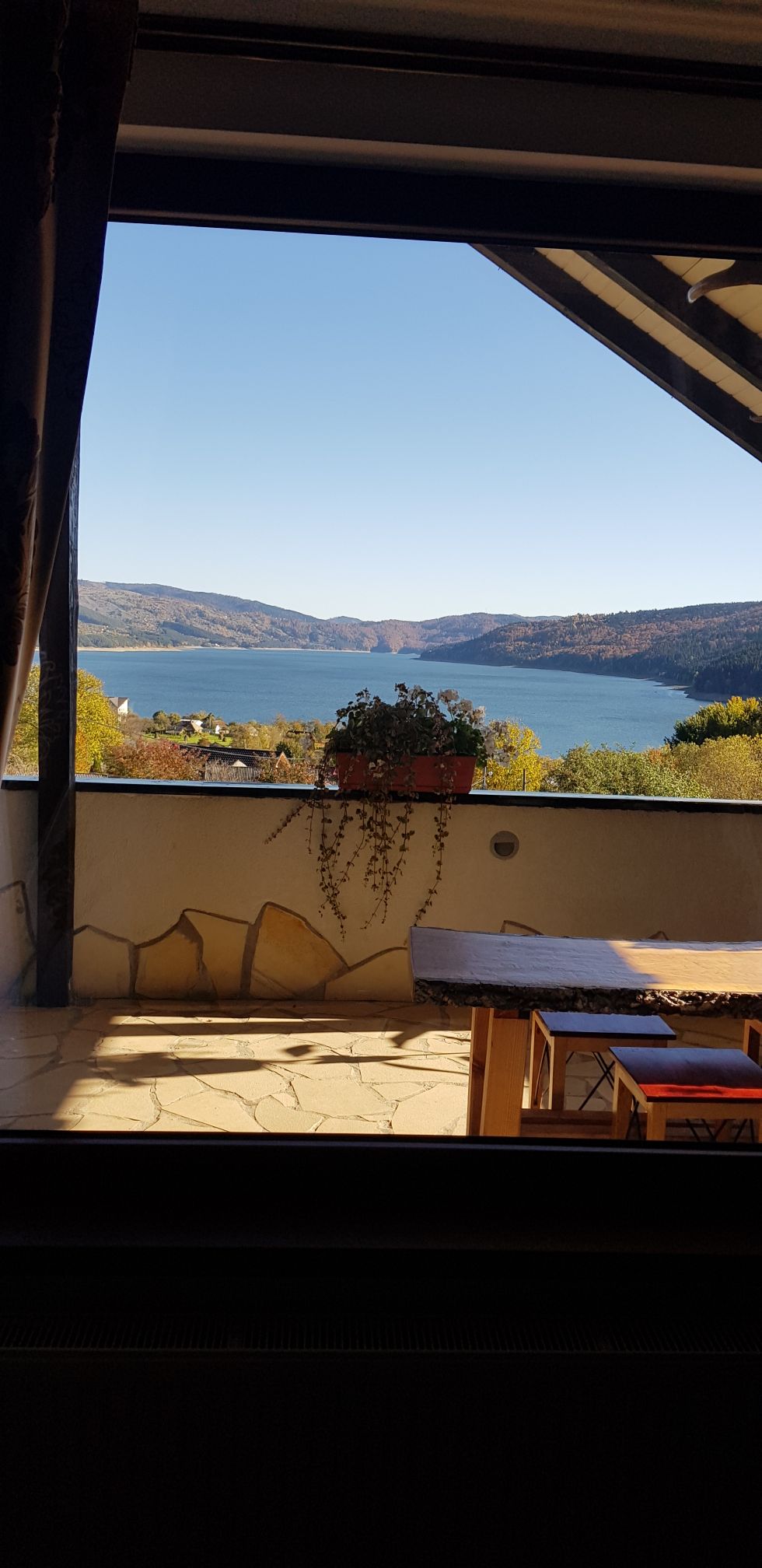 A scenic view is framed by a wide open window, showcasing a tranquil lake and rolling hills in the distance. A wooden table and chairs are positioned on a stone patio, inviting relaxation amidst the natural surroundings.