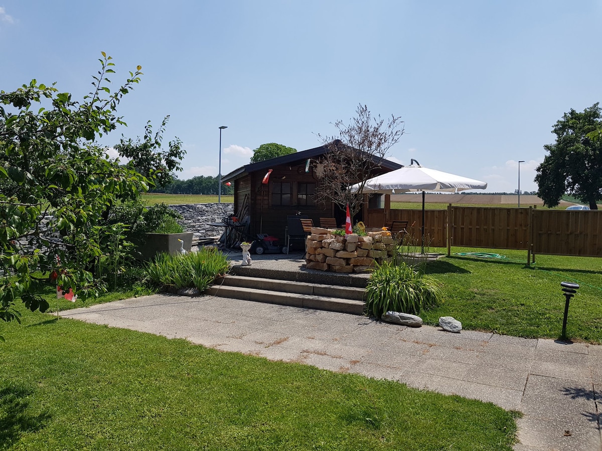 A small wooden chalet is nestled among greenery, featuring outdoor seating under a large umbrella. Stone steps lead up to the entrance, with a landscaped area of grass and flowerbeds surrounding it. The setting is calm, with a wooden fence and open fields visible in the background.