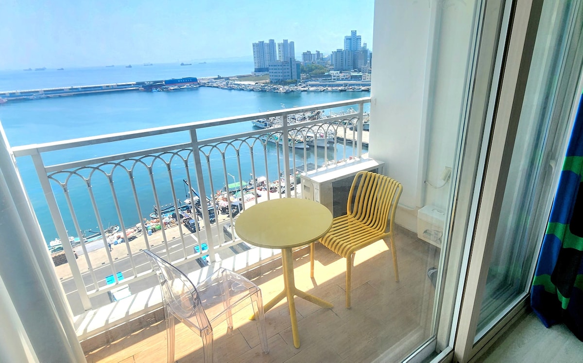 A balcony view showcases clear blue waters and a bustling harbor. A round yellow table and two transparent chairs are positioned on the balcony, framed by a glass railing that allows for unobstructed views of the surrounding landscape.