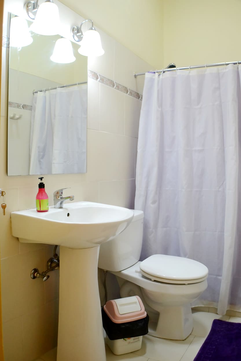 A clean and well-lit bathroom features a sink with a modern faucet, complemented by a large mirror above it. A white shower curtain is drawn to one side, revealing a shower area. The toilet is situated next to a waste bin.
