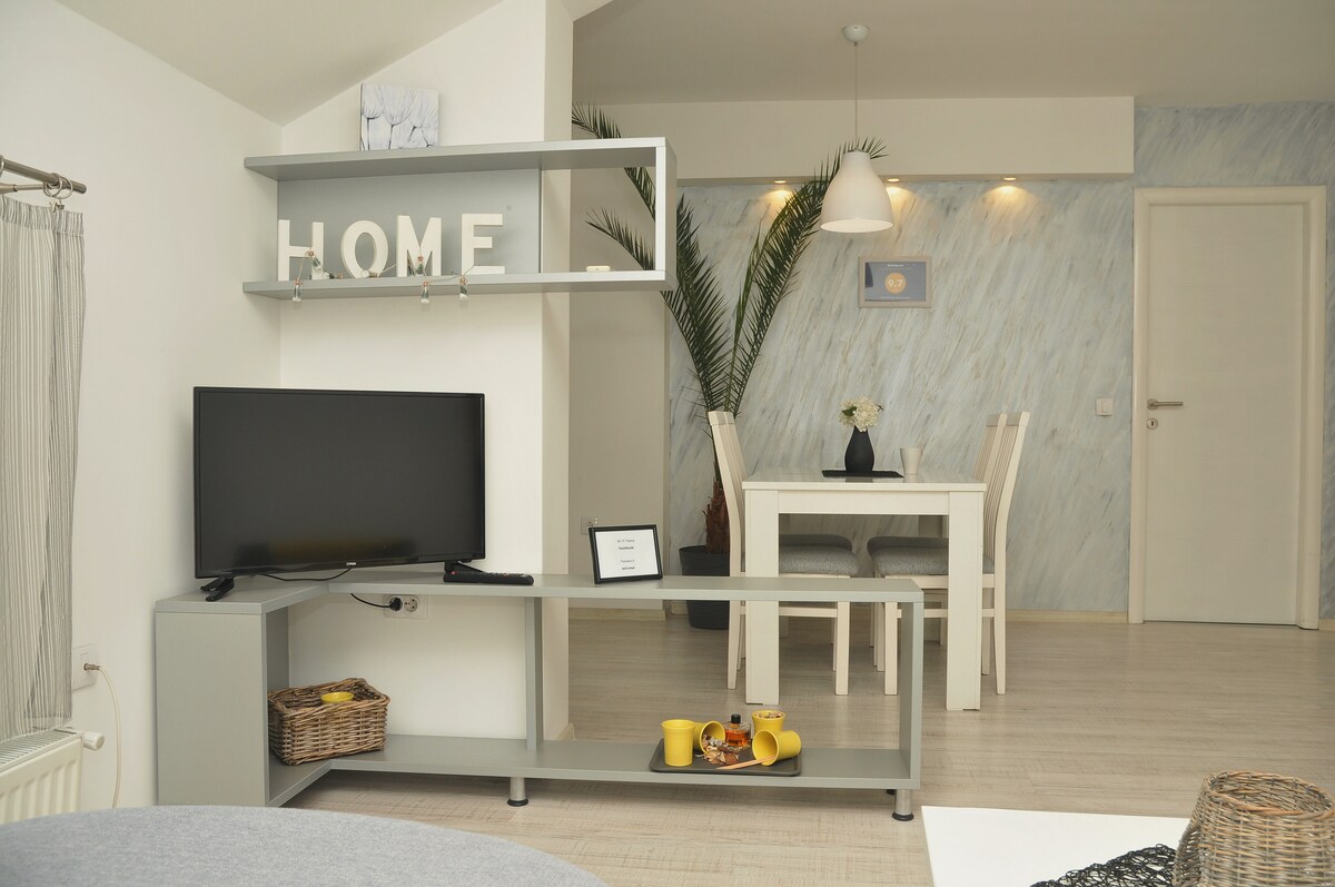 A modern living area is shown, featuring a flat-screen TV on a sleek console with decorative accents. A dining table with white chairs is visible in the background, complemented by a subtle wall design. A potted plant adds a touch of greenery.