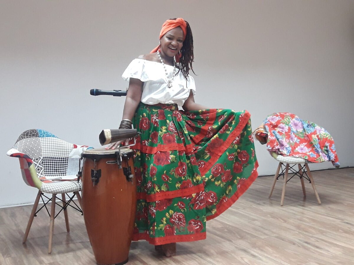 A performer is seen in a colorful skirt adorned with floral patterns, posing beside a traditional drum. Two chairs are positioned nearby, one draped with a vibrant fabric. The warm wooden floor complements the scene's inviting atmosphere.