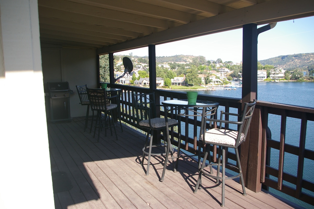 A spacious deck is showcased, featuring a dining set with black chairs and a table. A grill is visible in the background, and potted greenery adds a touch of color. The view overlooks the serene waterfront, providing a tranquil setting for relaxation.