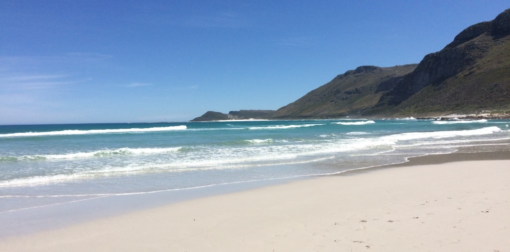 A serene beach scene features soft, white sand meeting gentle waves under a clear blue sky. Rolling hills rise in the background, adding depth to the natural landscape. The horizon blends seamlessly with the ocean, creating a calm and inviting atmosphere.