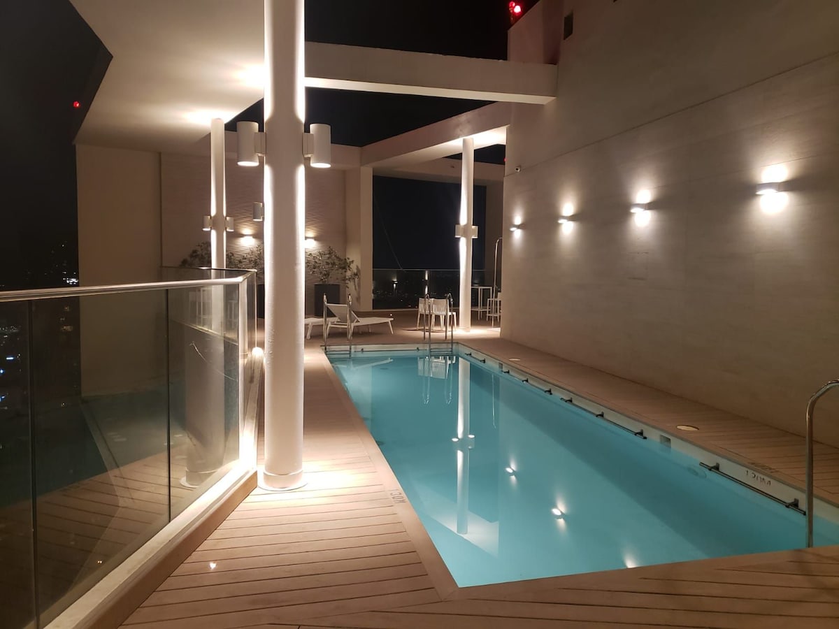 A rooftop pool area is highlighted in this image, featuring a clear blue swimming pool surrounded by wood decking. Contemporary lounge chairs are positioned nearby, with soft lighting illuminating the space. Glass railings provide an unobstructed view of the surrounding cityscape at night.