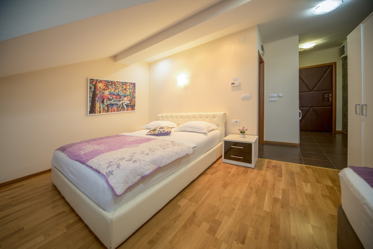 A spacious bedroom is presented with a light-colored bed featuring a tufted headboard. Decorative artwork adorns the wall above the bed. A small nightstand holds a plant, while wooden flooring enhances the warm atmosphere. Natural light fills the room through the window, creating an inviting space.