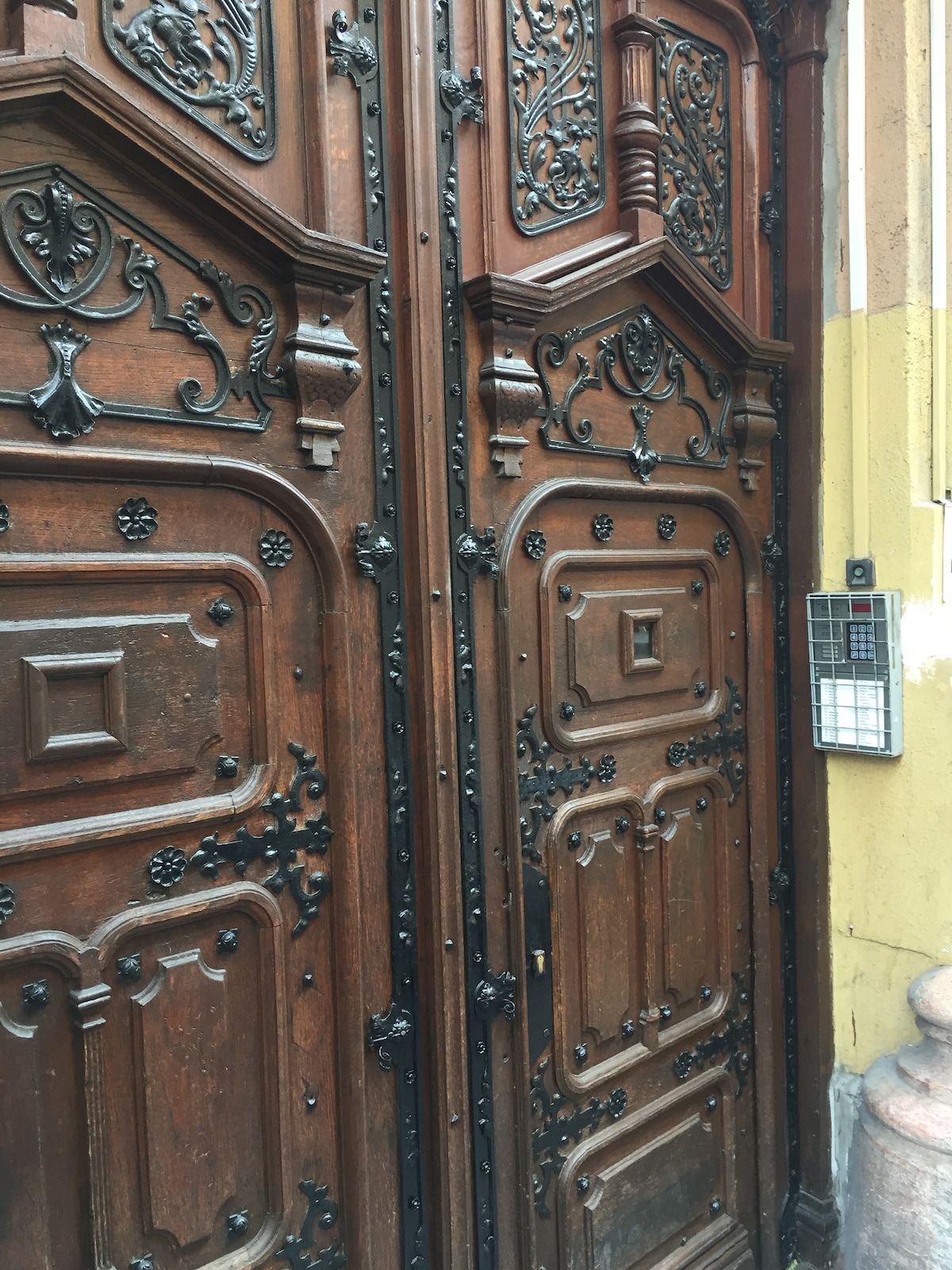 A pair of ornate wooden doors is displayed, featuring intricate carvings and a dark, glossy finish. Decorative black metal accents enhance the design. A small intercom box is positioned to the right, mounted on the warm-colored wall beside the doors.