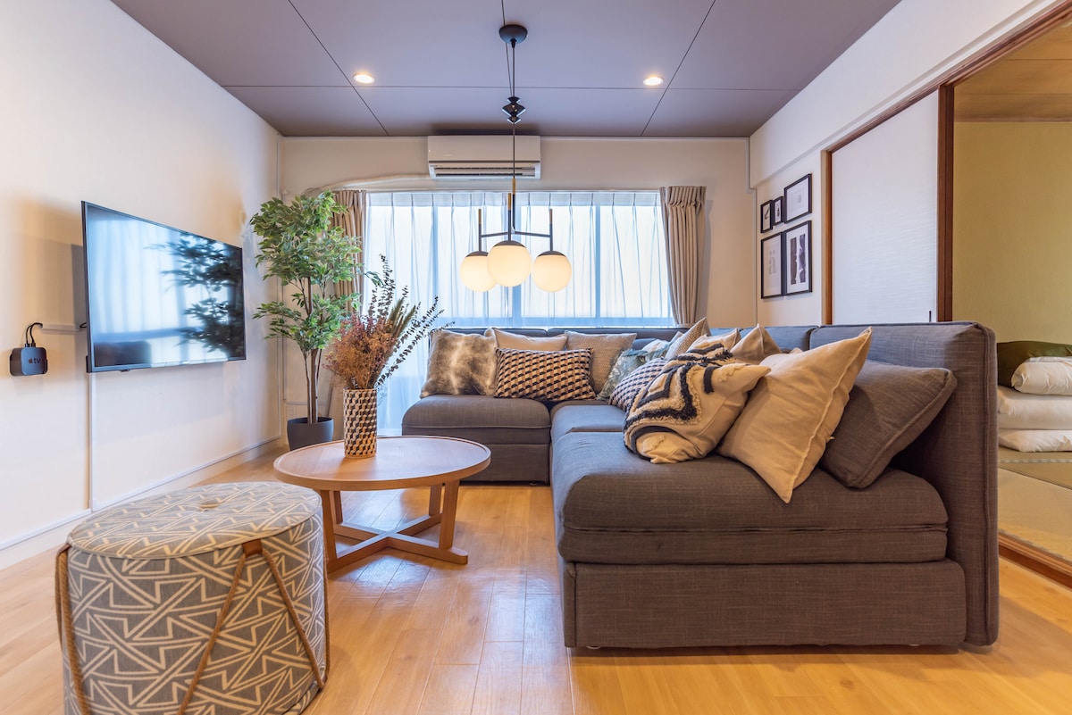 The living area features a sectional sofa adorned with decorative pillows, positioned near a light wood coffee table. A television is mounted on the wall opposite the seating, while natural light filters through the large window, complemented by a plant in the corner.