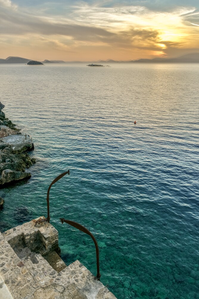 The image captures a serene seascape during sunset, with gentle waves lapping at the rocky shore. Two rustic handrails descend into the calm water, while distant islands are visible against the horizon, accentuated by vibrant hues of orange and blue in the sky.