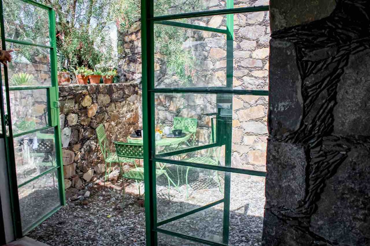 A green metal door leads to a private outdoor space, showcasing a small table and chairs surrounded by stone walls. Potted plants add a natural touch to the earthy setting, with sunlight filtering through the leafy foliage.