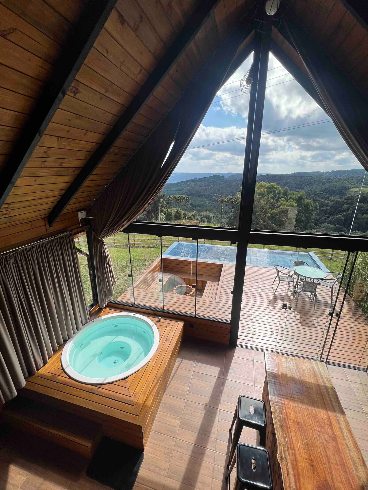 An internal area showcases a wooden hot tub, surrounded by floor-to-ceiling glass walls. Natural light fills the space, revealing a stunning view of the outdoors. A wooden deck, featuring a table and chairs, leads to an inviting heated pool, accentuating the relaxing atmosphere.