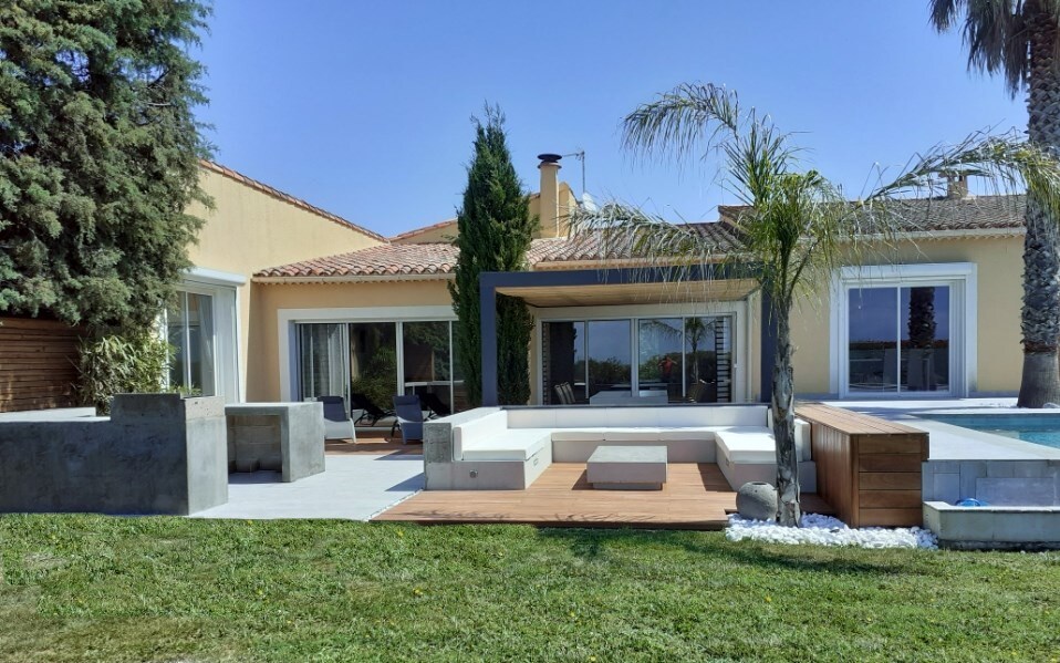 The exterior of the villa features a modern design with large glass doors, providing a seamless connection to the outdoor space. A stylish lounge area is created with a combination of wooden and stone materials, surrounded by a well-maintained garden and palm trees.