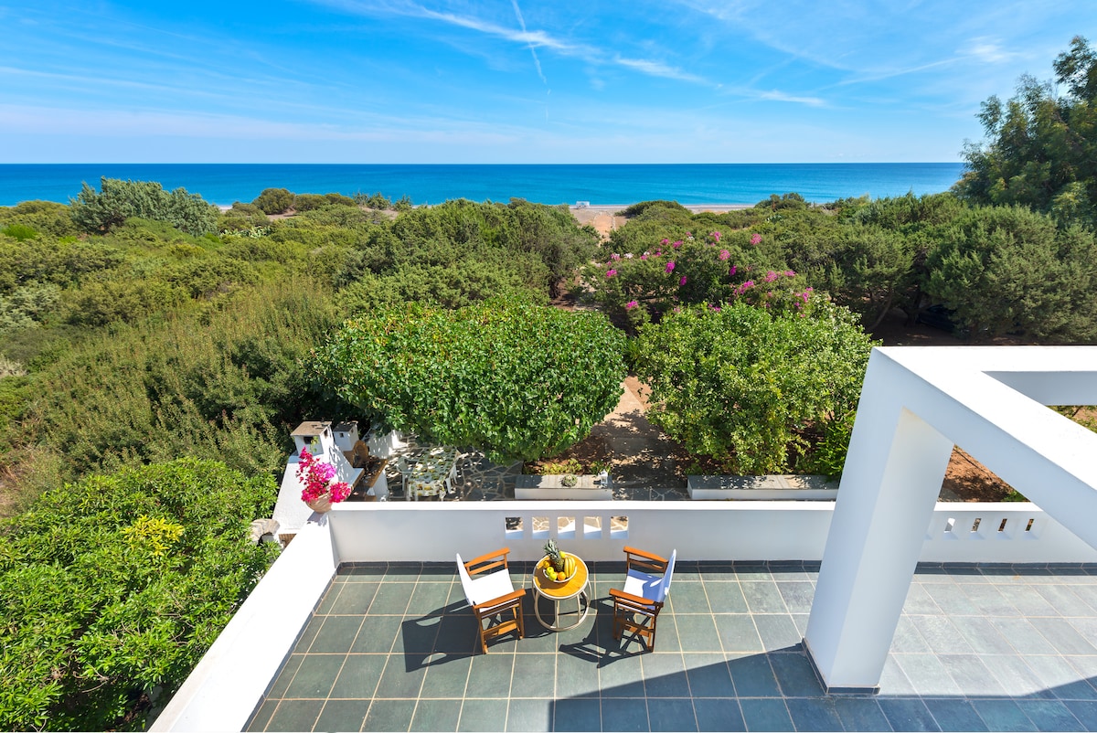 A spacious terrace is presented, featuring two wooden chairs and a small table. Lush greenery surrounds the area, offering a view of the sparkling sea in the distance. The blue sky complements the vibrant natural setting, creating a serene outdoor space.