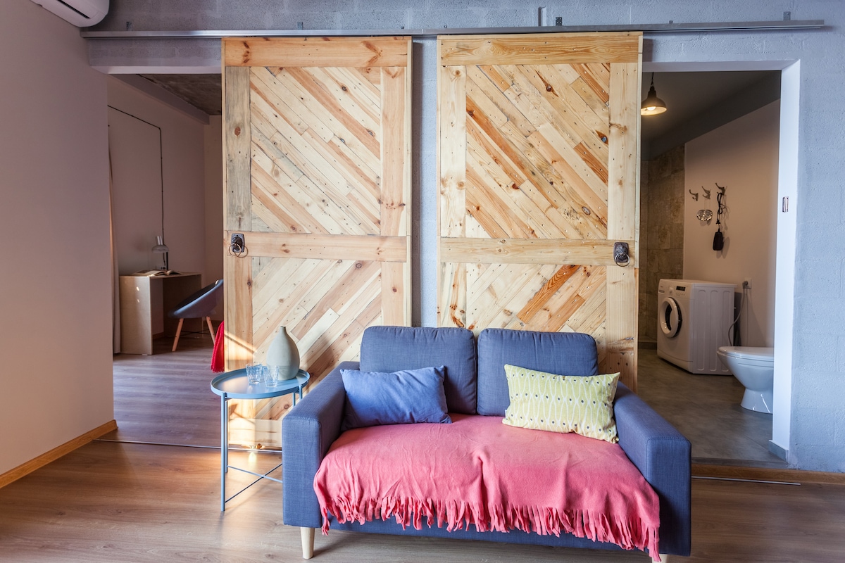 A modern living area features a gray sofa adorned with a colorful throw and decorative pillows. Behind it, striking wooden doors with a diagonal pattern provide access to a separate space. The room is warmly lit, offering a welcoming and comfortable atmosphere.