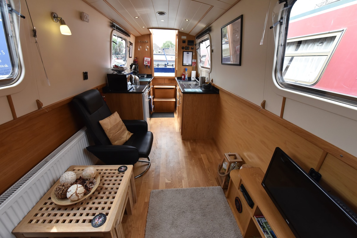 A cozy living space features wooden paneling and a warm wooden floor. A black recliner chair is positioned beside a small table with a plate of snacks. A compact kitchen area is visible at the back, complemented by light from windows that frame the canal view.