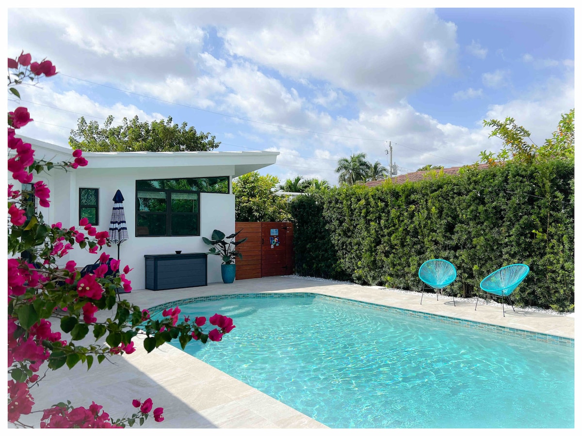 A sparkling saltwater pool is surrounded by lush greenery, providing a refreshing retreat. Two turquoise chairs are placed near the water's edge, inviting relaxation. The vibrant pink flowers frame the scene, adding a touch of color to the tranquil atmosphere.