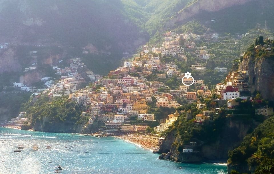 A scenic view of Positano’s colorful buildings cascading along the hillside, overlooking the blue waters of the beach. The lush greenery of the mountains frames the town, creating a striking coastal landscape. The image captures the blend of natural beauty and architectural charm of the area.