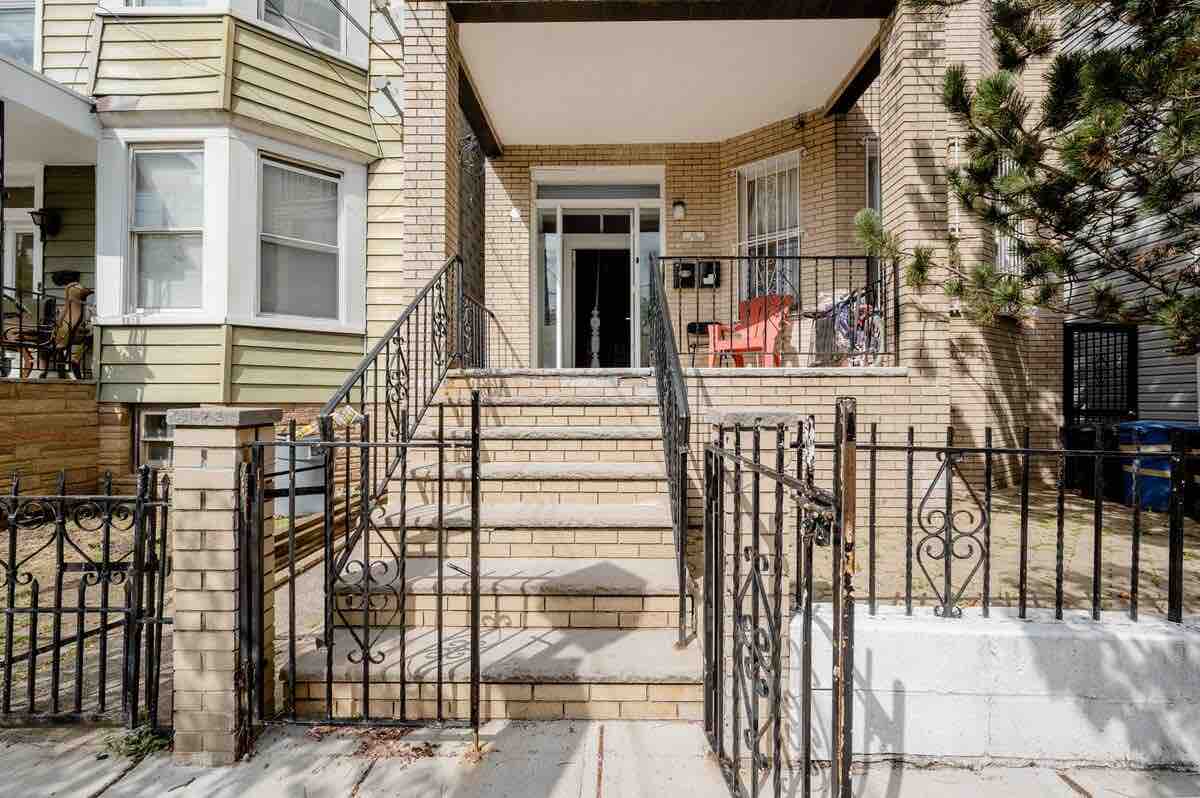 An inviting entrance features a well-maintained staircase leading to the front door, framed by decorative railings. The building's facade is composed of light-colored bricks, accentuated by large windows. Bright red chairs add a pop of color to the porch area, creating a welcoming first impression.