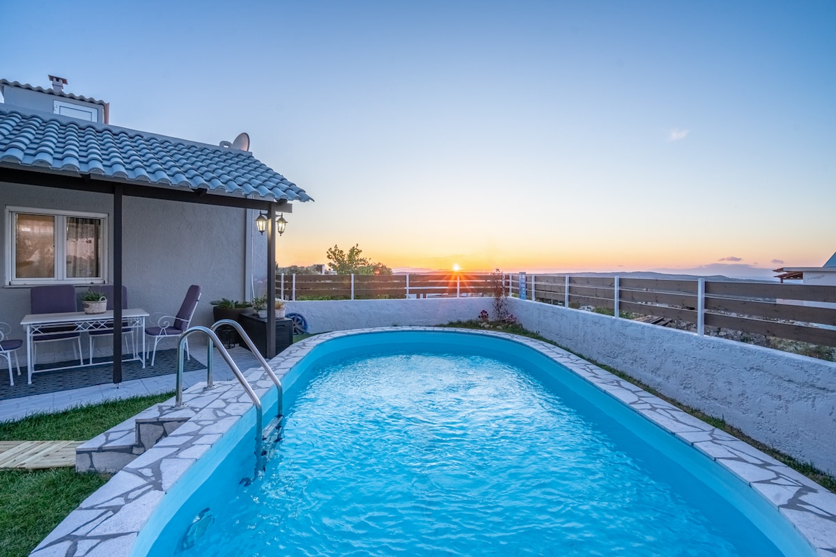 A private eco pool is surrounded by a neatly paved area, with a glimpse of a dining space featuring a table and chairs nearby. The sunset casts warm colors across the horizon, creating a serene backdrop against the white fence and rustic mountain scenery.