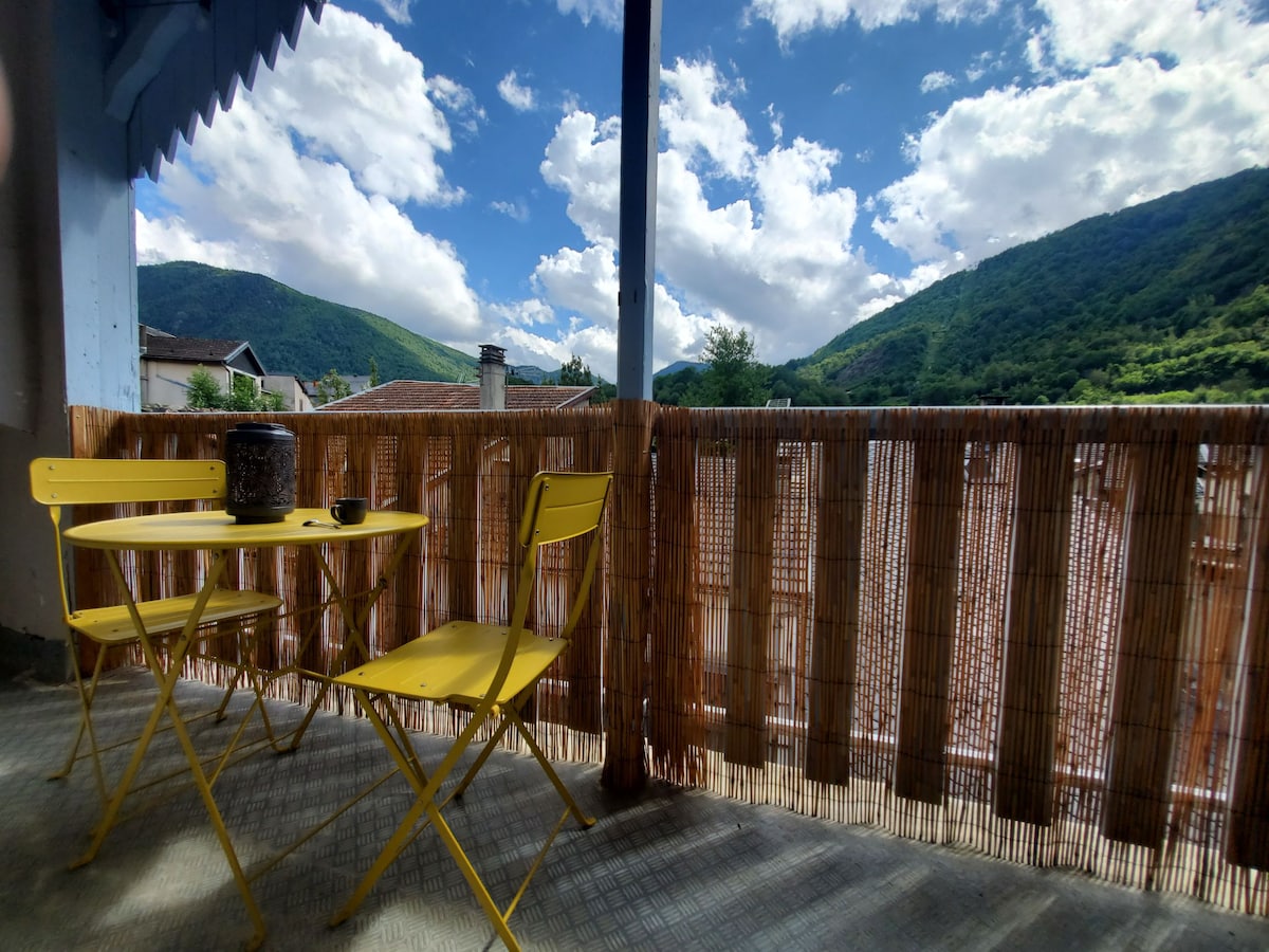 A small terrace features a round yellow table accompanied by two folding chairs, offering a tranquil space to enjoy outdoor views. The surrounding fence showcases a natural bamboo design, framing a scenic backdrop of green mountains and a partly cloudy blue sky.