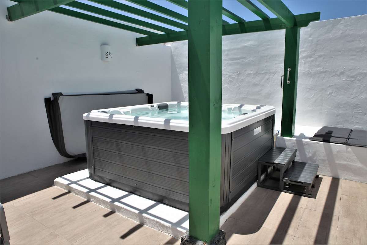 A private Jacuzzi for five people is situated under a green pergola, surrounded by white walls. Steps provide easy access, while the smooth tile flooring ensures a safe area for relaxation. Natural light highlights the design, promoting an inviting space for unwinding.