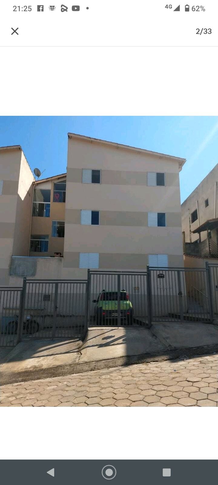 The exterior of a two-story building is visible, featuring a light-colored facade and multiple windows. A gated entrance leads to the parking area, where a small green car is parked. The clear blue sky enhances the scene's brightness.