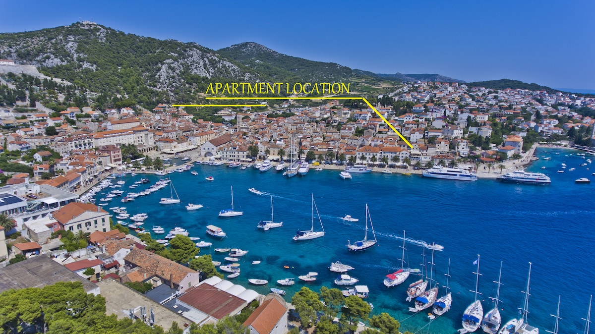 An aerial view captures the scenic landscape of Hvar, featuring a vibrant harbor filled with boats and lined by picturesque buildings. Lush green hills rise in the background, and the apartment location is clearly marked, showcasing proximity to the waterfront and town center.