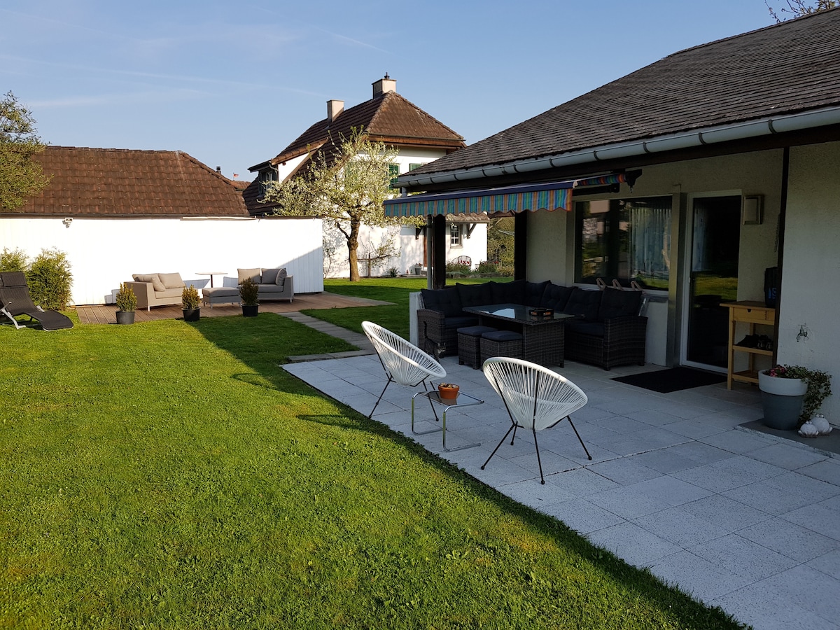 A spacious garden area is visible, featuring a paved patio with seating, including two armchairs and a sectional sofa. Lush green grass surrounds the seating area, and a striped awning provides shade. The setting is quiet and serene, with a clear sky in the background.