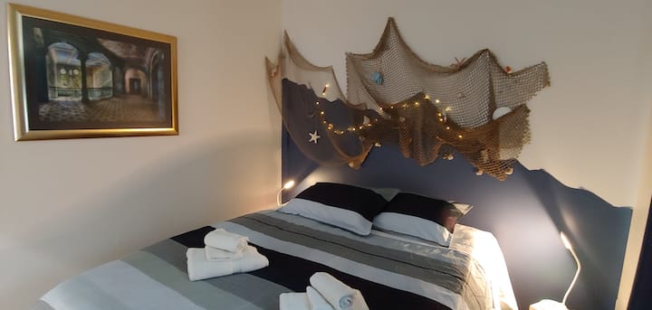 Una camera da letto con design ispirato all'oceano, con asciugamani sul letto e comodini con lampade che offrono luci rosa, blu e verdi. Sulle pareti ci sono opere d'arte e una rete da pesca con reperti oceanici.