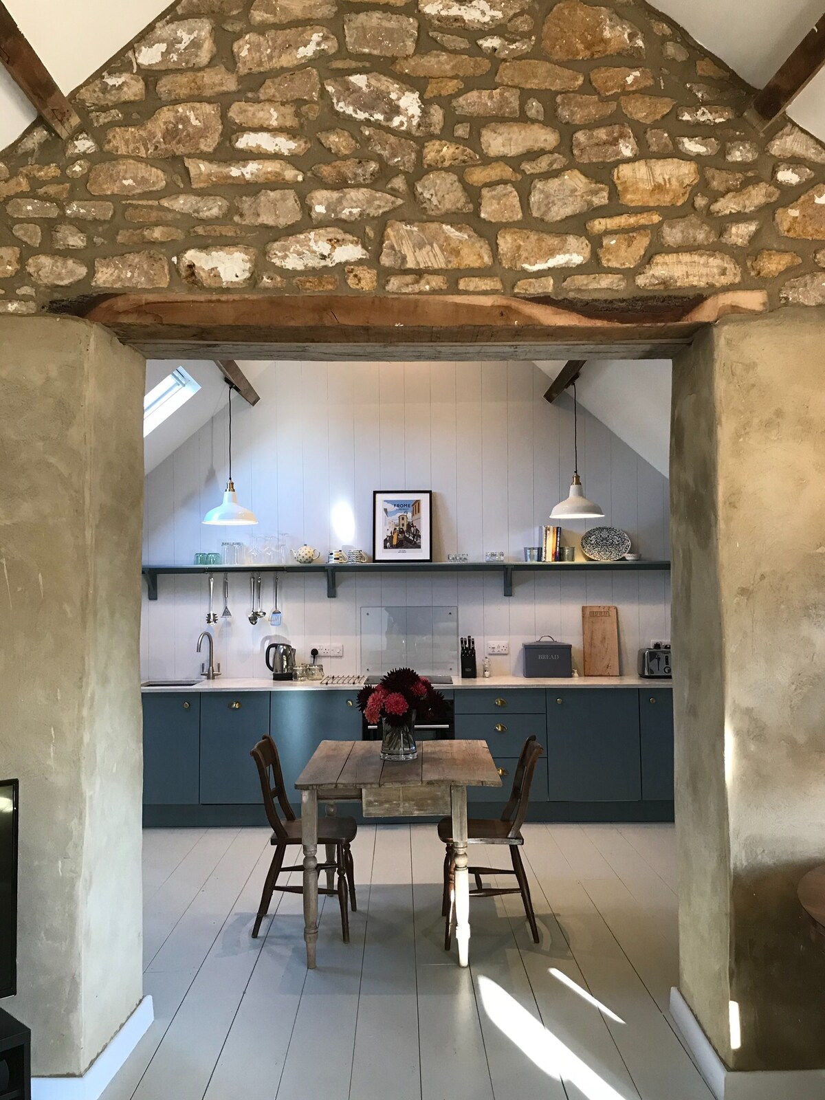 A spacious kitchen area featuring exposed stone walls and wooden beams creates a rustic feel. A small wooden table with two chairs is positioned in the foreground. The kitchen is well-equipped with modern appliances, and decorative items are displayed on the shelves above.