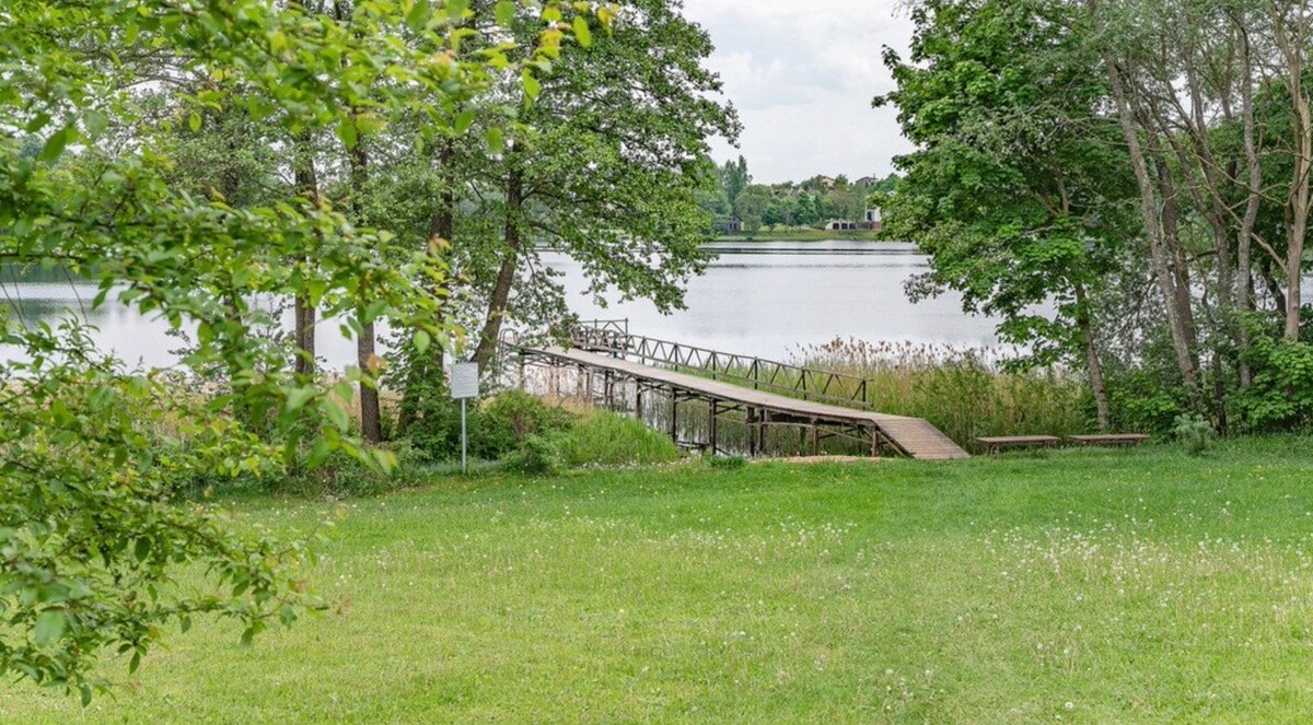 A serene view of a wooden dock extends into a calm lake, framed by lush greenery. The grassy area in the foreground features scattered wildflowers, inviting outdoor relaxation. Soft reflections of trees are visible on the water's surface, contributing to the peaceful atmosphere.