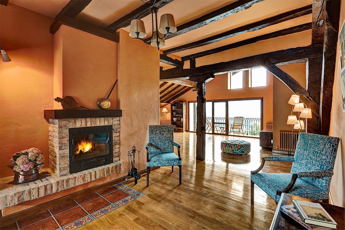 A spacious living area features exposed wooden beams and warm-colored walls. A cozy fireplace with a brick surround adds warmth, while two turquoise chairs are positioned nearby. Natural light fills the space through large windows, revealing a view of the outdoors.