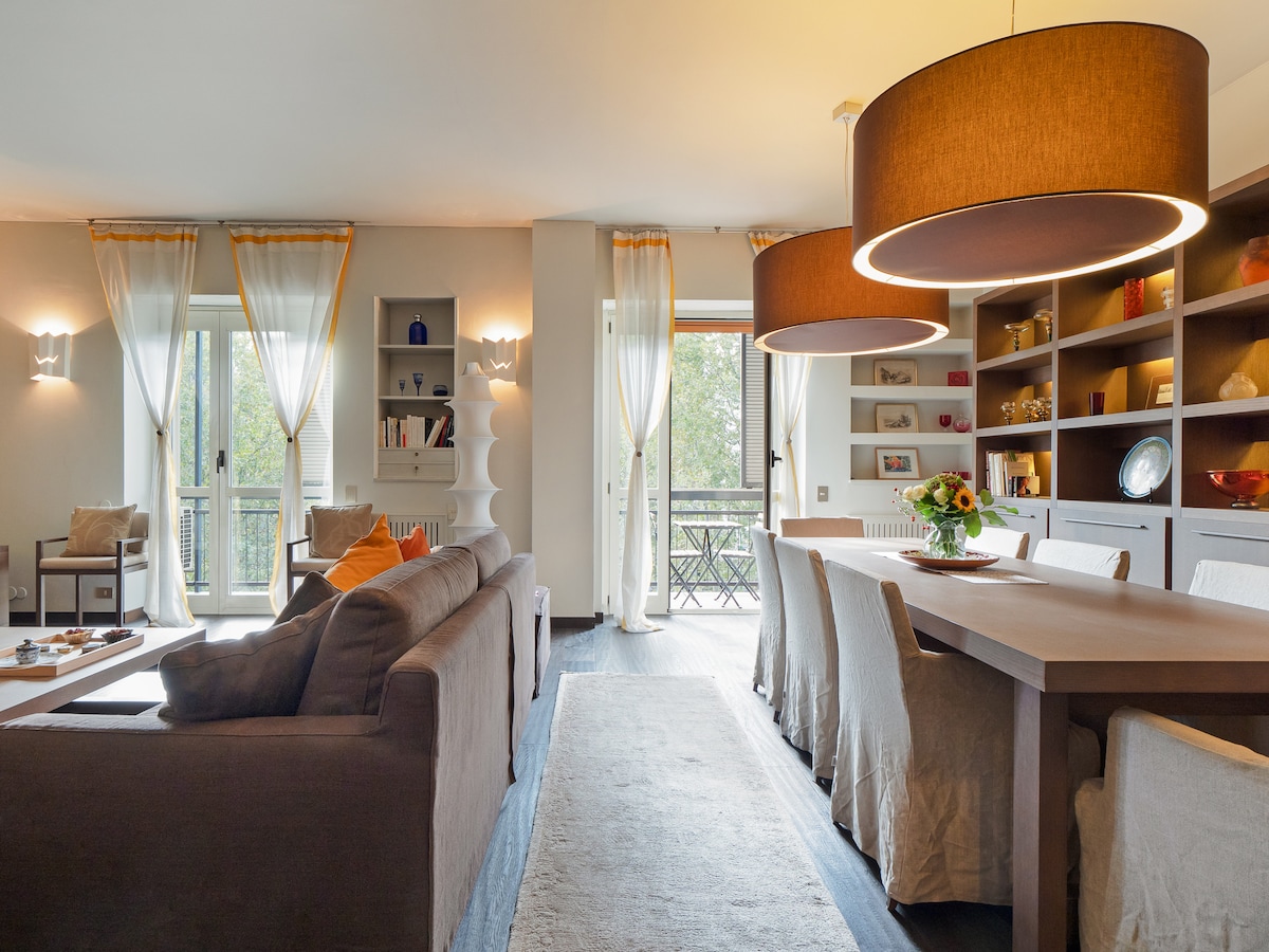 A spacious living area includes a comfortable sofa and dining table, with large pendant lights above. Two sets of glass doors open to a balcony, filling the space with natural light. Shelving units display decorative items, enhancing the room's inviting ambience.