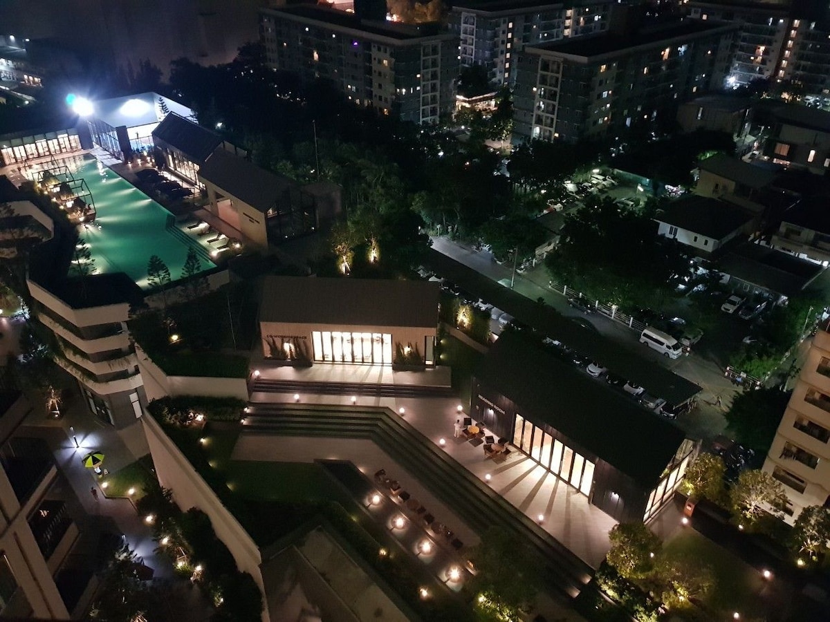 A nighttime view displays the well-lit grounds of the condo complex, highlighting various amenities such as the swimming pool and surrounding greenery. Residential buildings and pathways are visible, with cars parked nearby, creating a sense of community and accessibility.